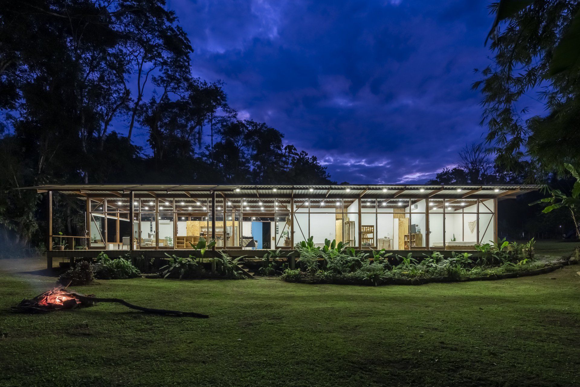 Uma grande casa de vidro é iluminada à noite com uma fogueira na frente.