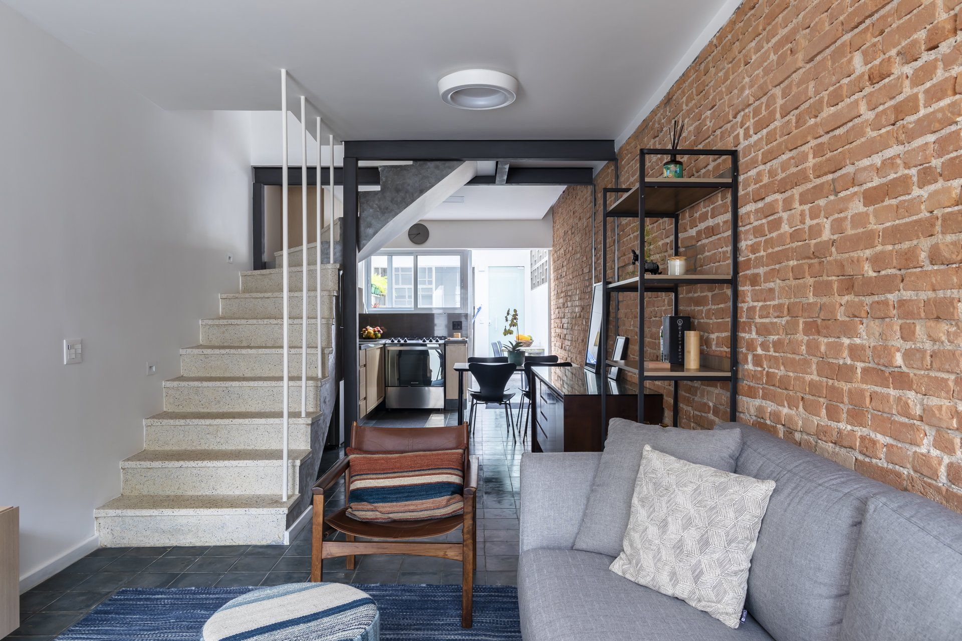 Uma sala com sofá, cadeira, escadas e parede de tijolos.