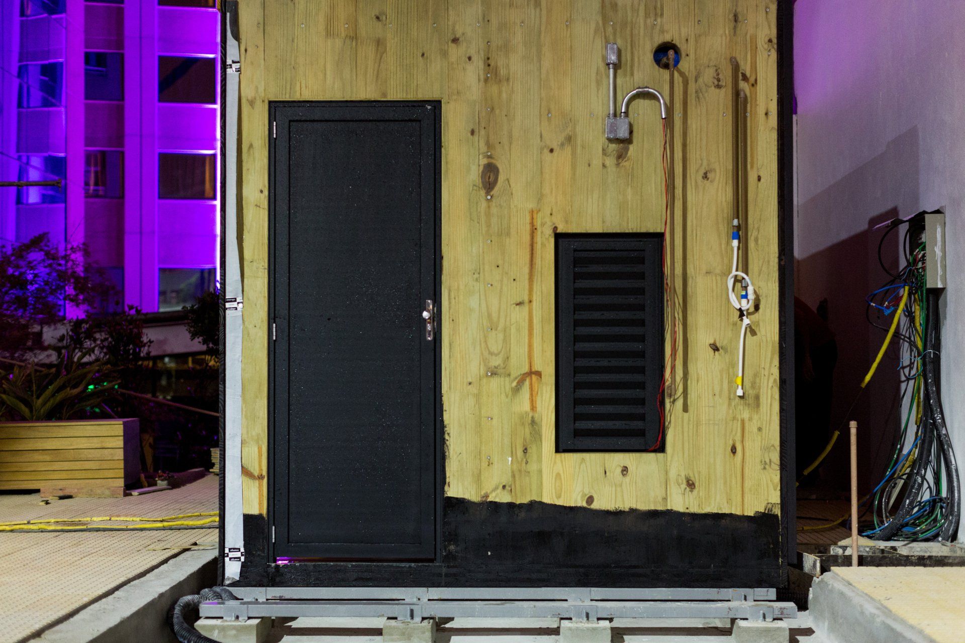 Um pequeno prédio de madeira com uma porta preta e um prédio roxo ao fundo.