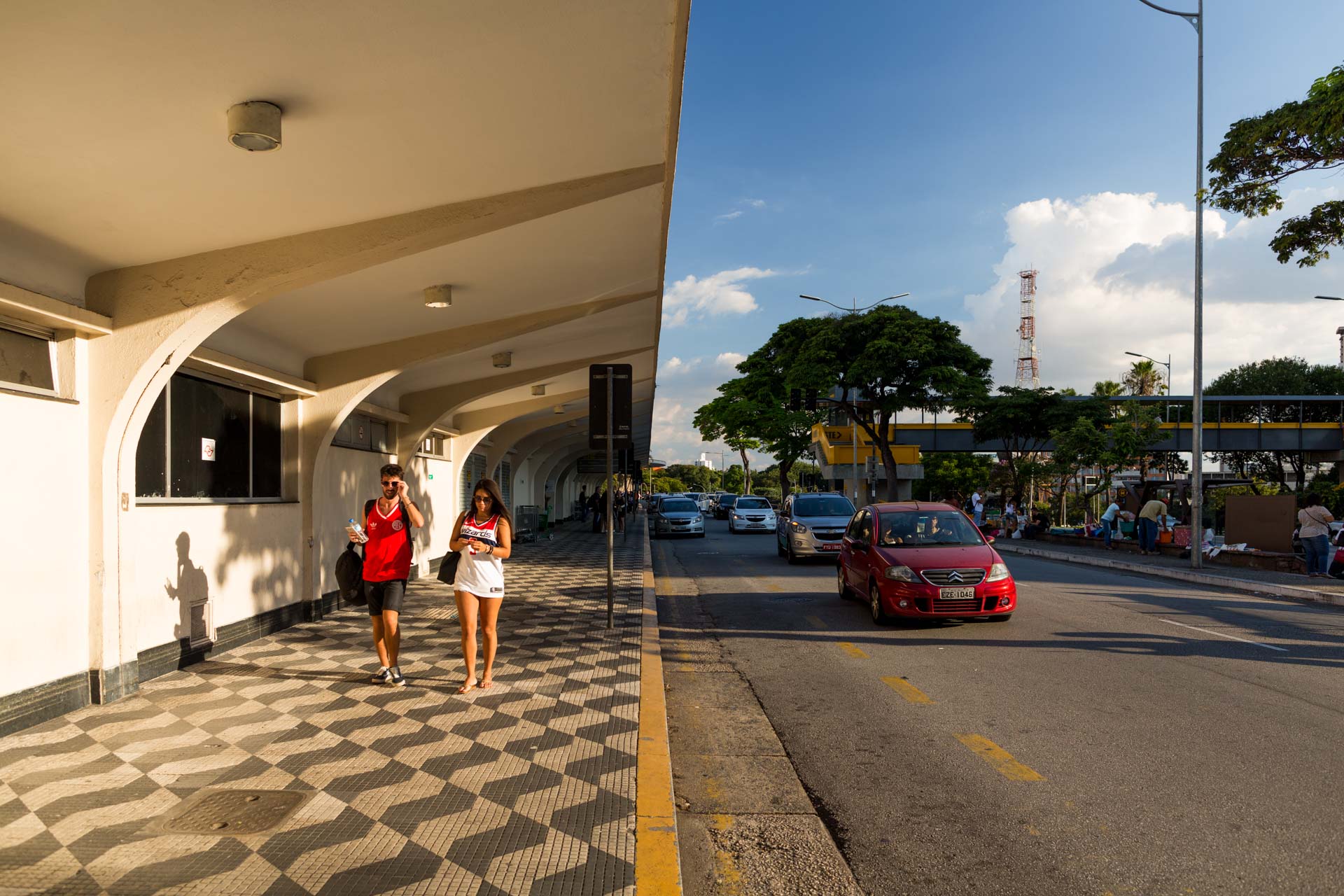 Algumas pessoas estão andando na calçada ao lado de um carro vermelho.