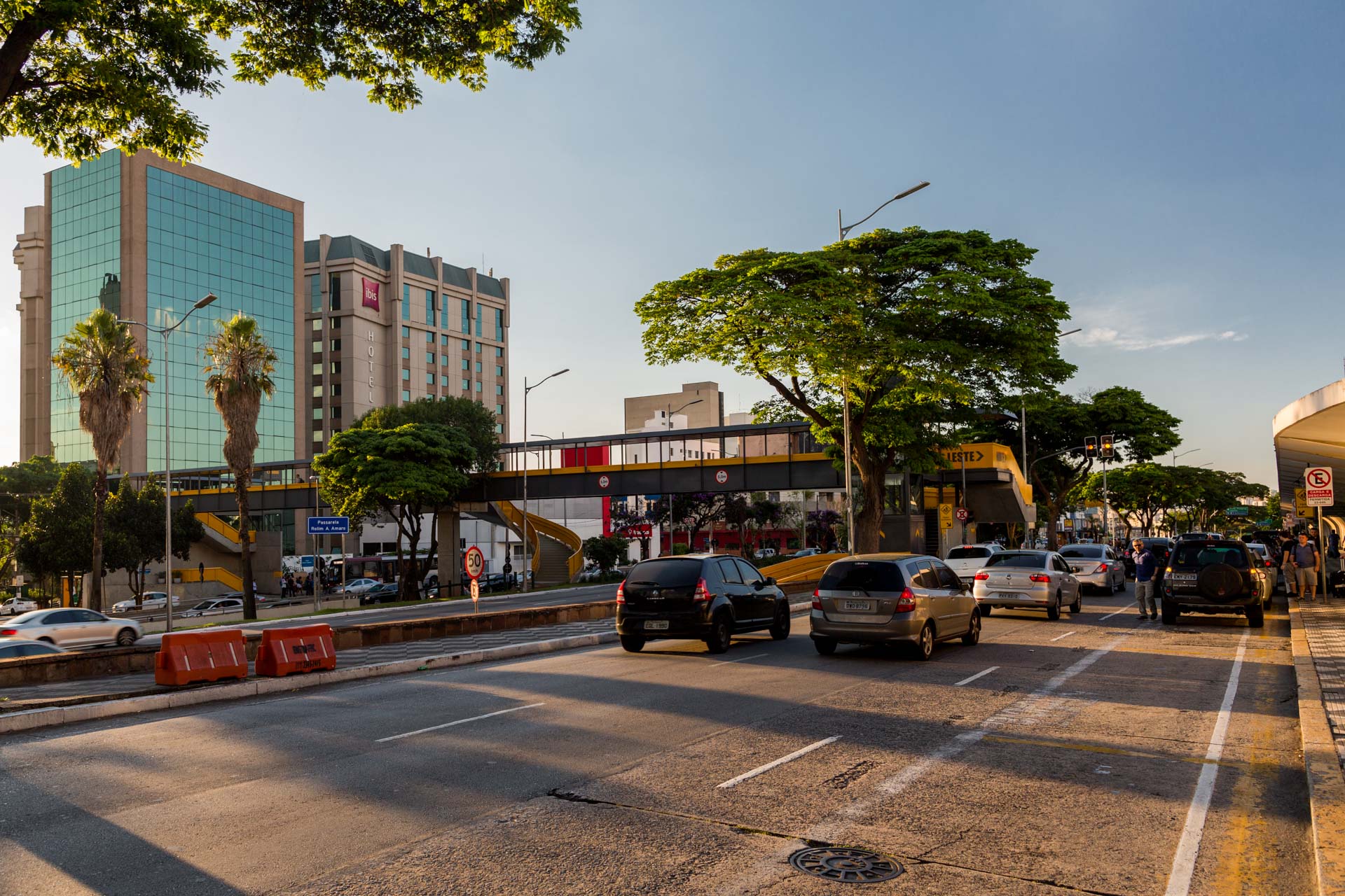 Uma rua movimentada da cidade com carros passando por ela e um prédio alto ao fundo.