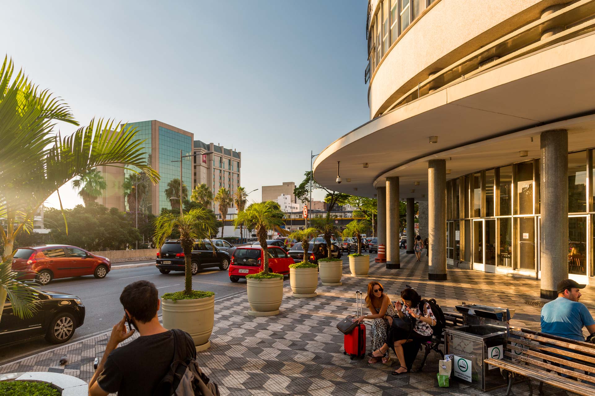 Um homem está falando ao celular enquanto está sentado em um banco em frente a um prédio.