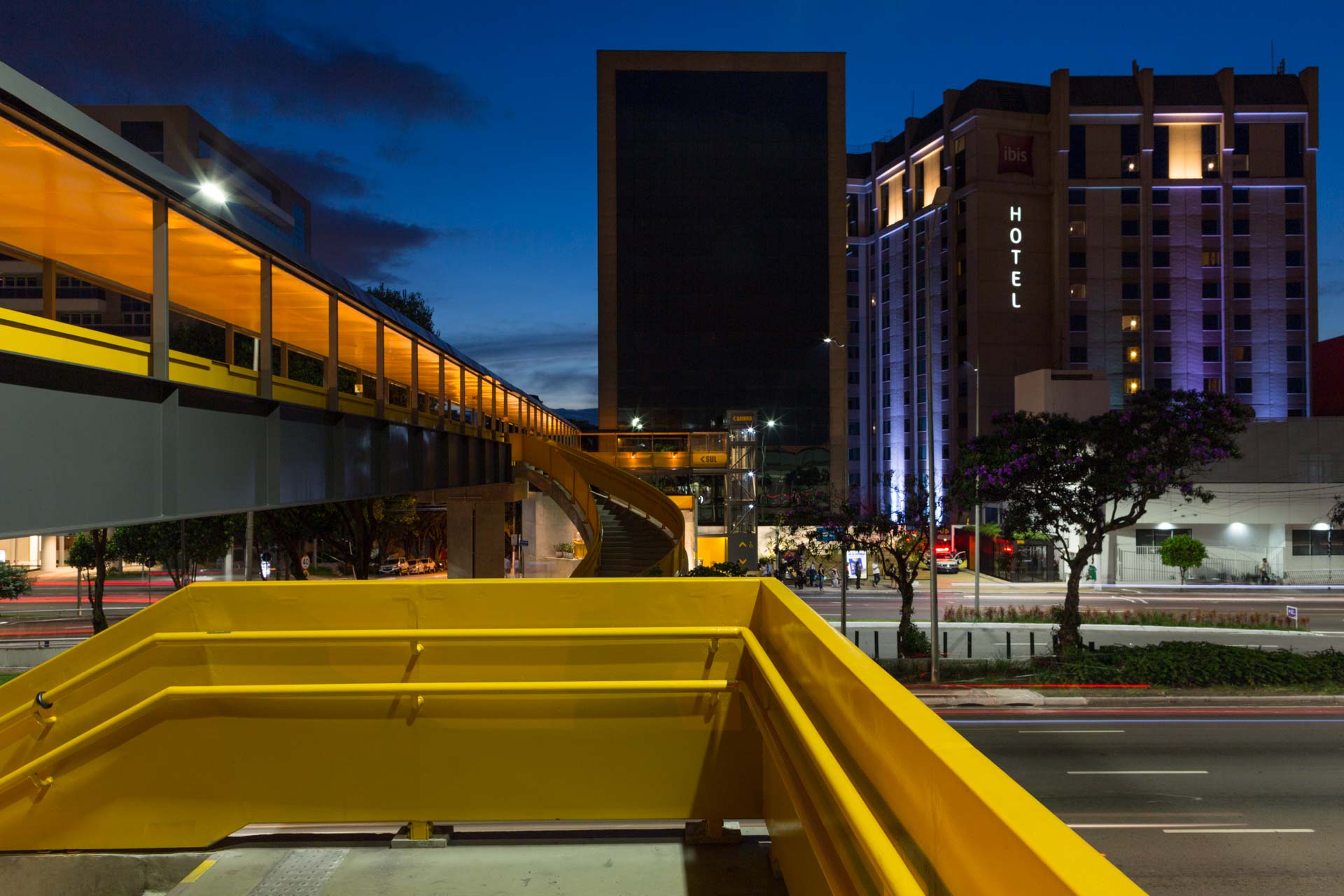 Uma ponte amarela sobre uma rua da cidade à noite com um hotel ao fundo.
