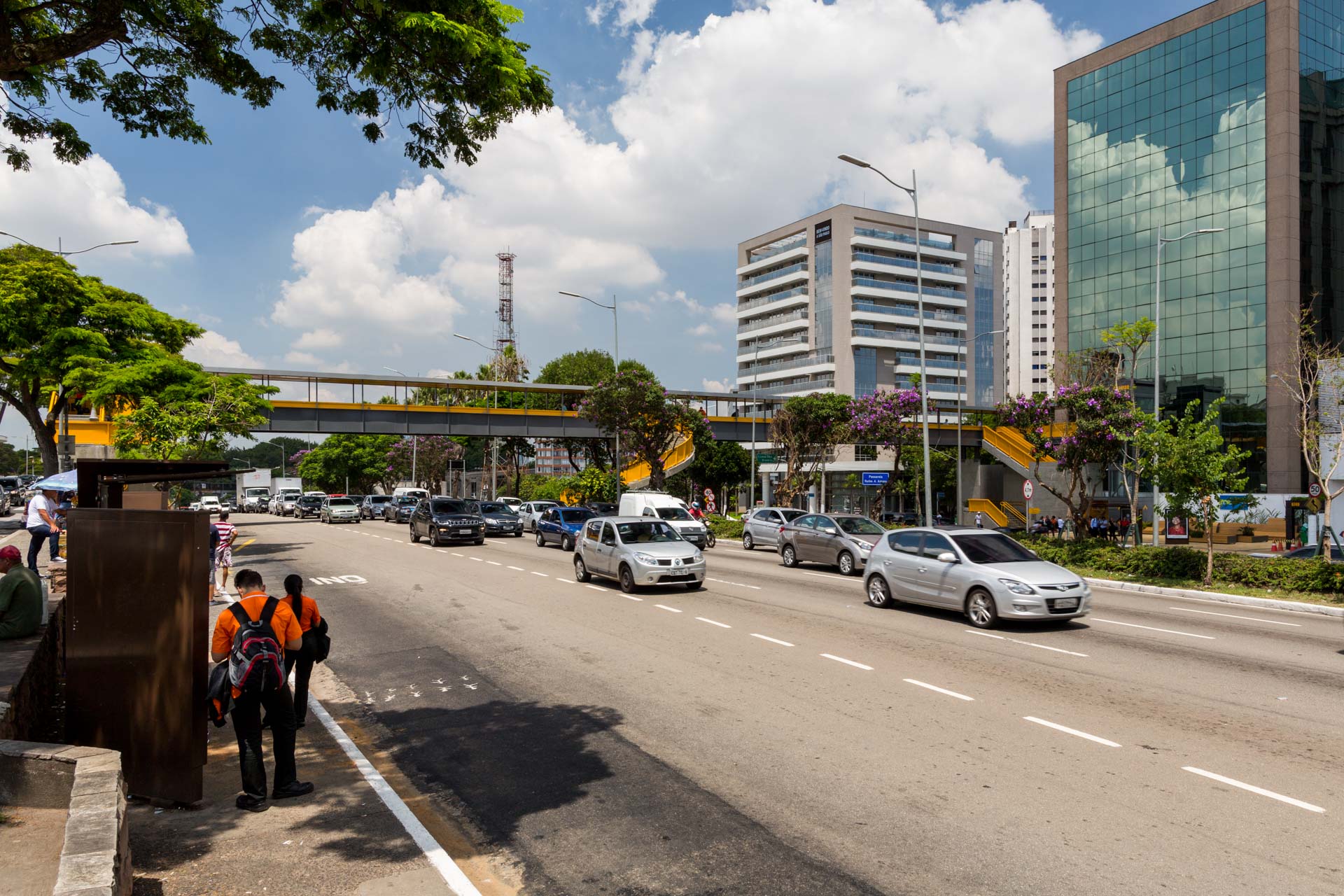Algumas pessoas estão paradas em um ponto de ônibus na beira de uma rua da cidade.
