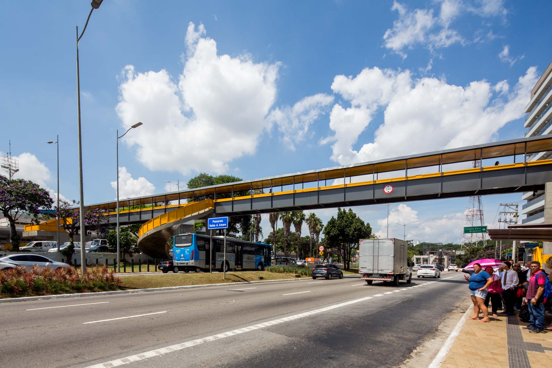 Um grupo de pessoas está esperando em um ponto de ônibus debaixo de uma ponte.