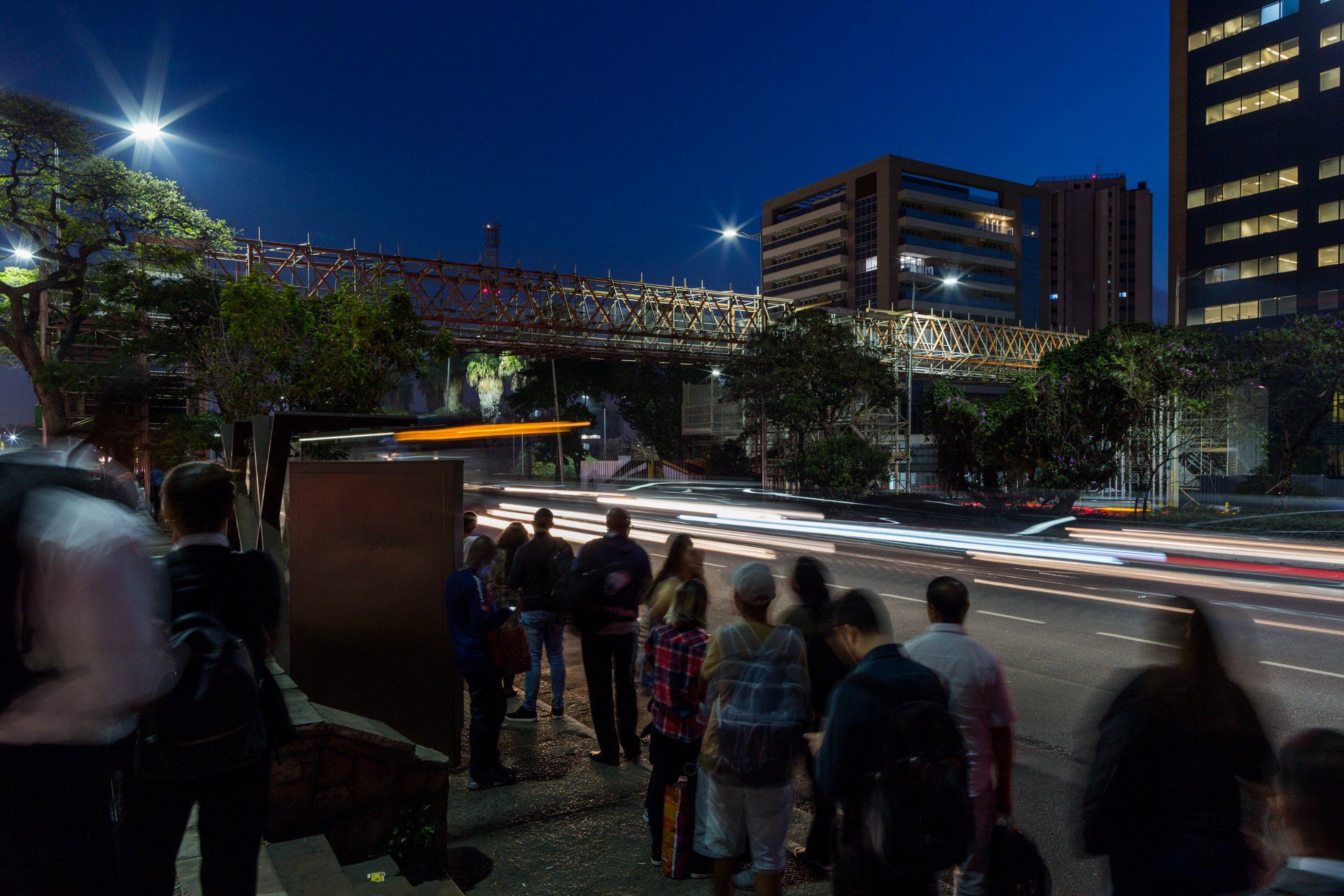Um grupo de pessoas está parado na beira de uma rua da cidade à noite.