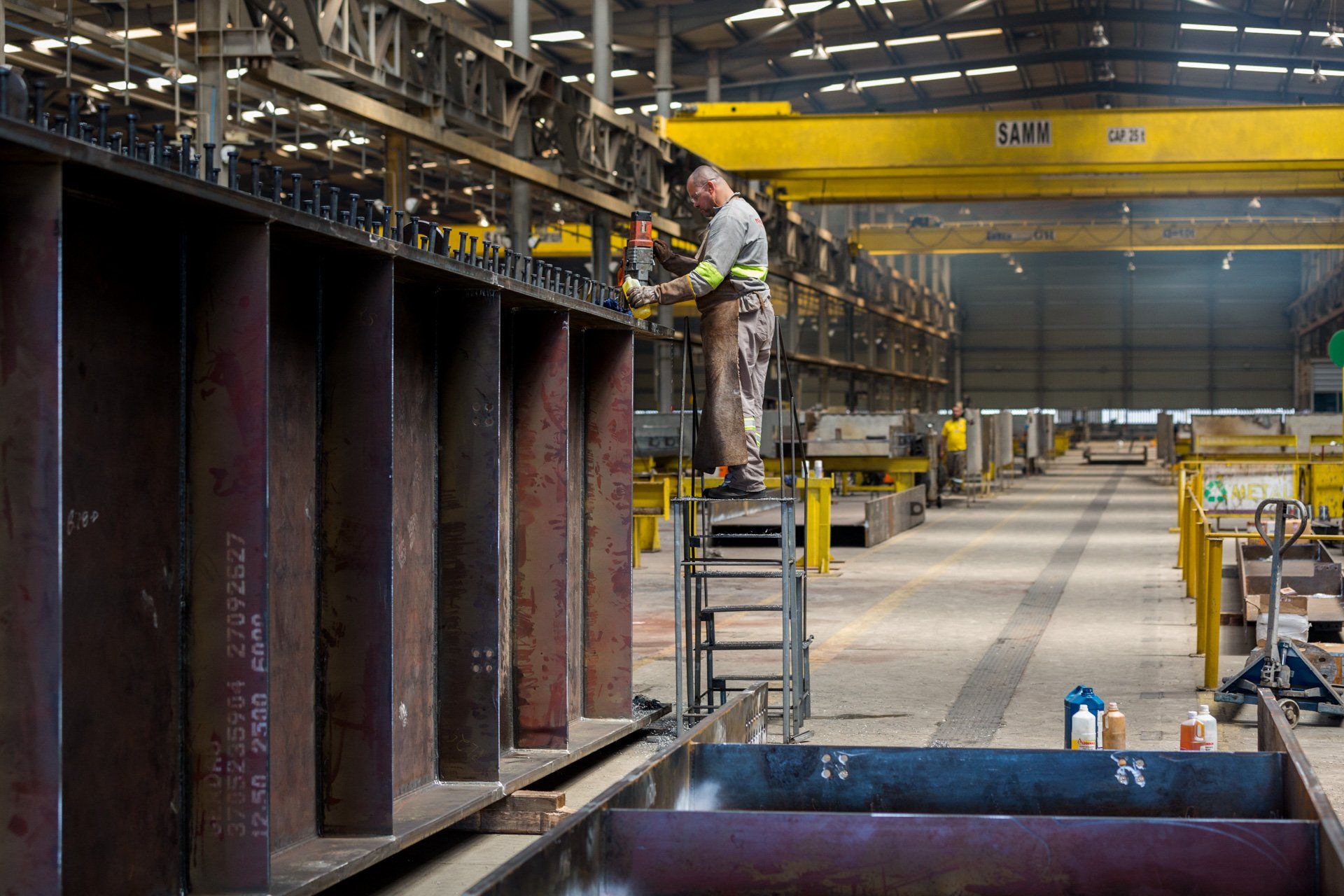 Um homem está em uma escada em uma fábrica trabalhando em uma estrutura metálica.