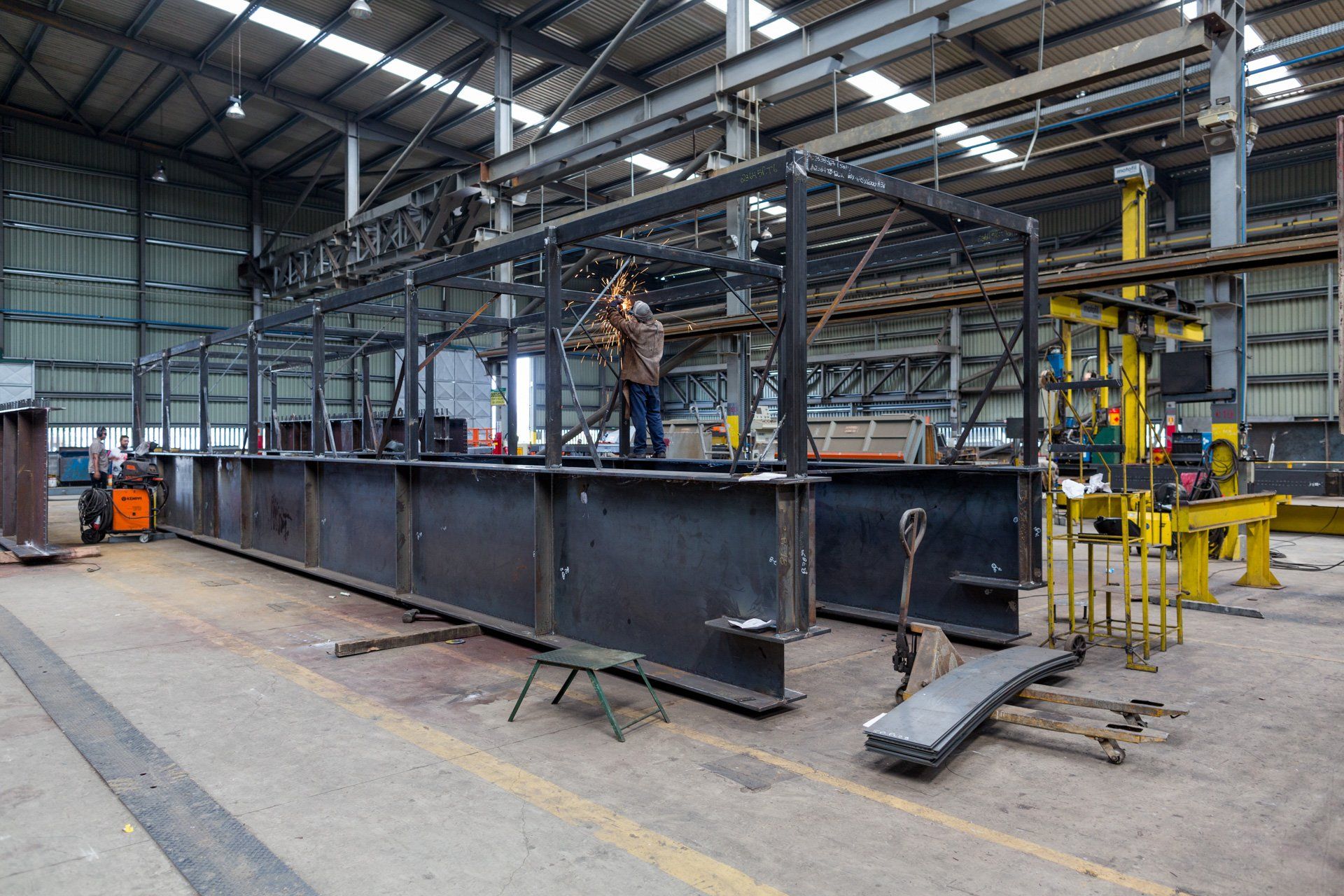 Um homem está soldando uma grande estrutura metálica em uma fábrica.