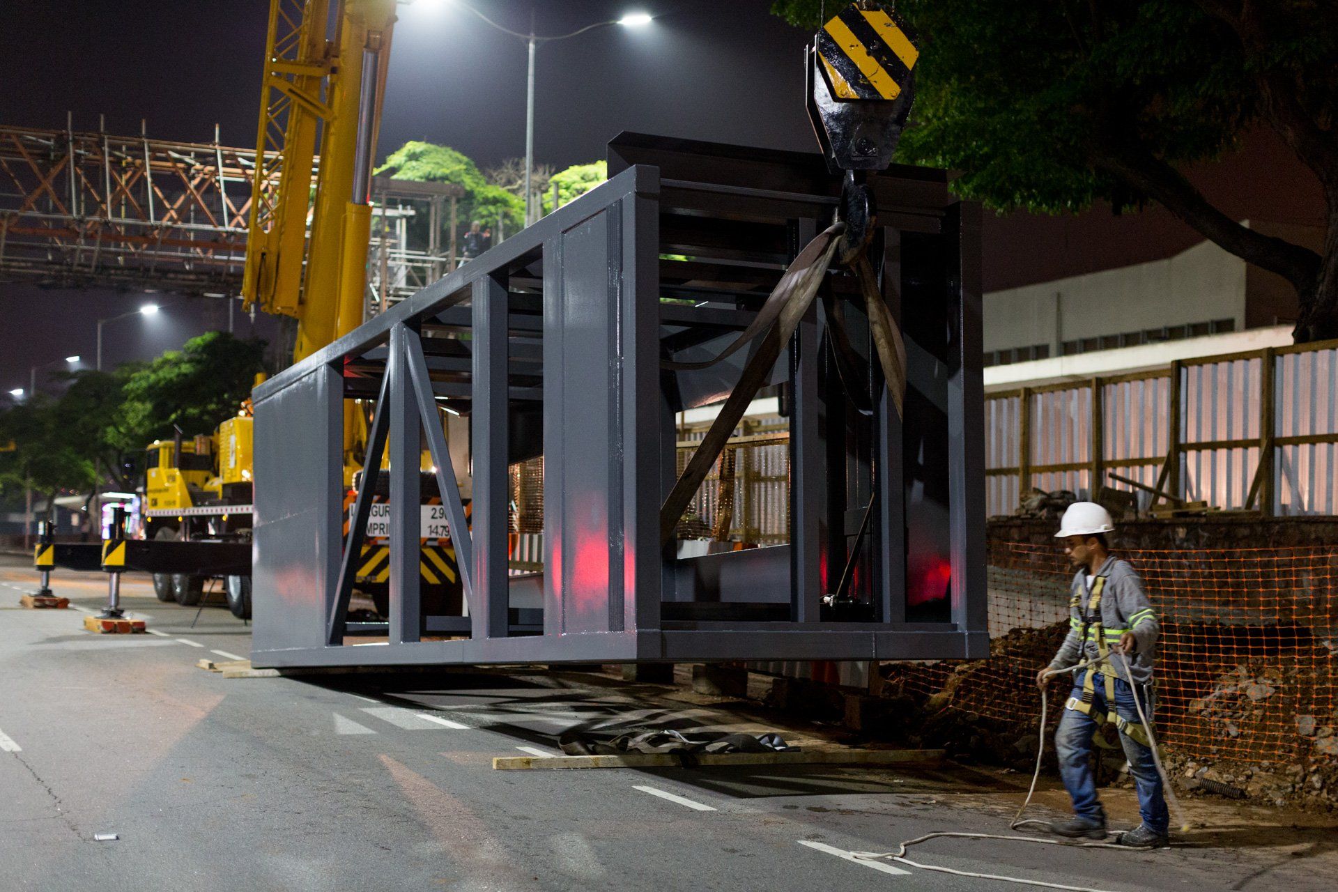 Um trabalhador da construção civil está parado na beira da estrada, próximo a uma grande estrutura metálica que está sendo levantada por um guindaste.