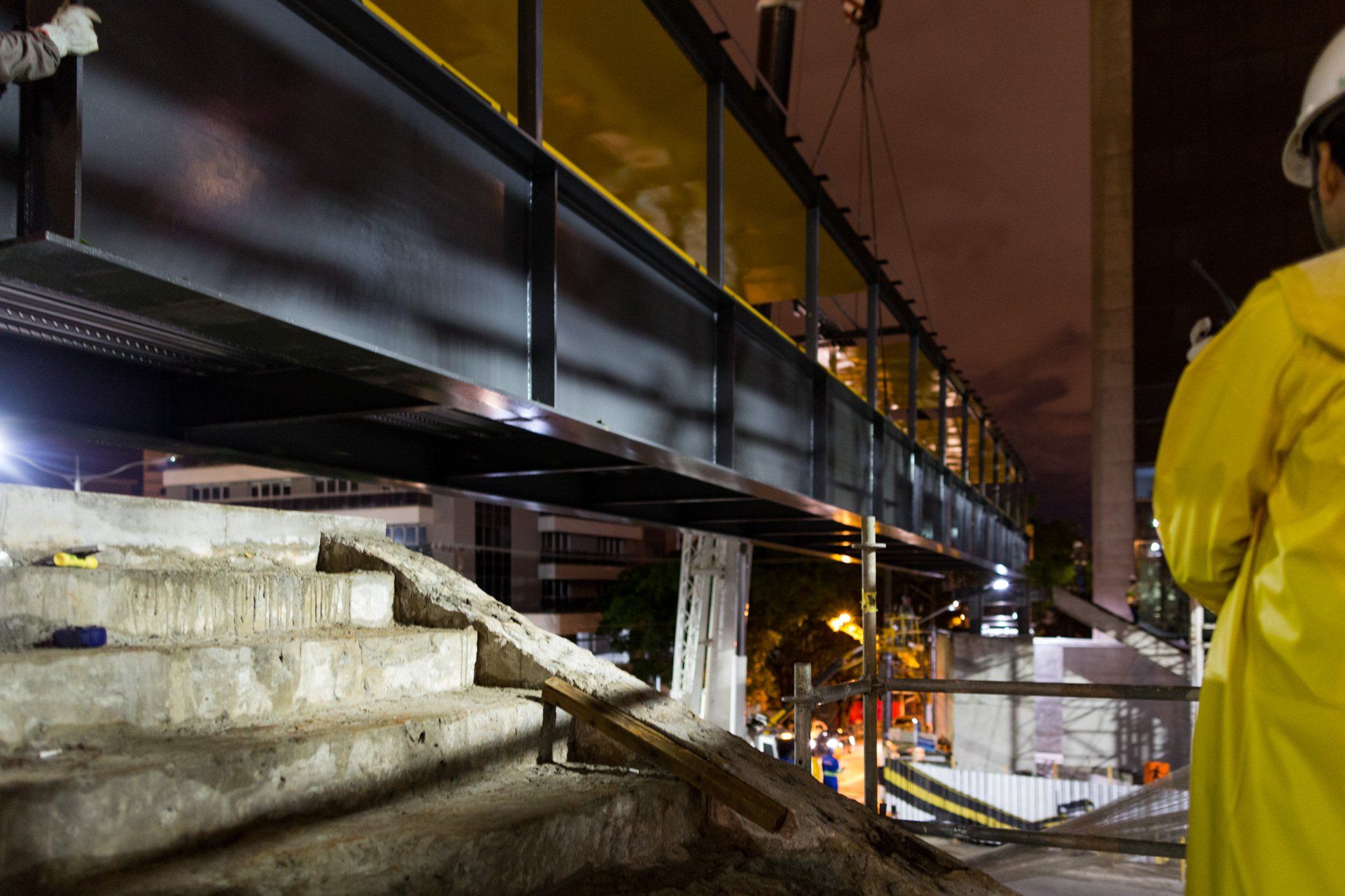 Um homem com uma capa de chuva amarela está parado em uma ponte à noite.