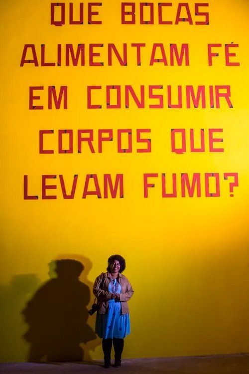 A woman stands in front of a yellow wall that says que bocas alimentam fe em consumir corpos que levam fumo