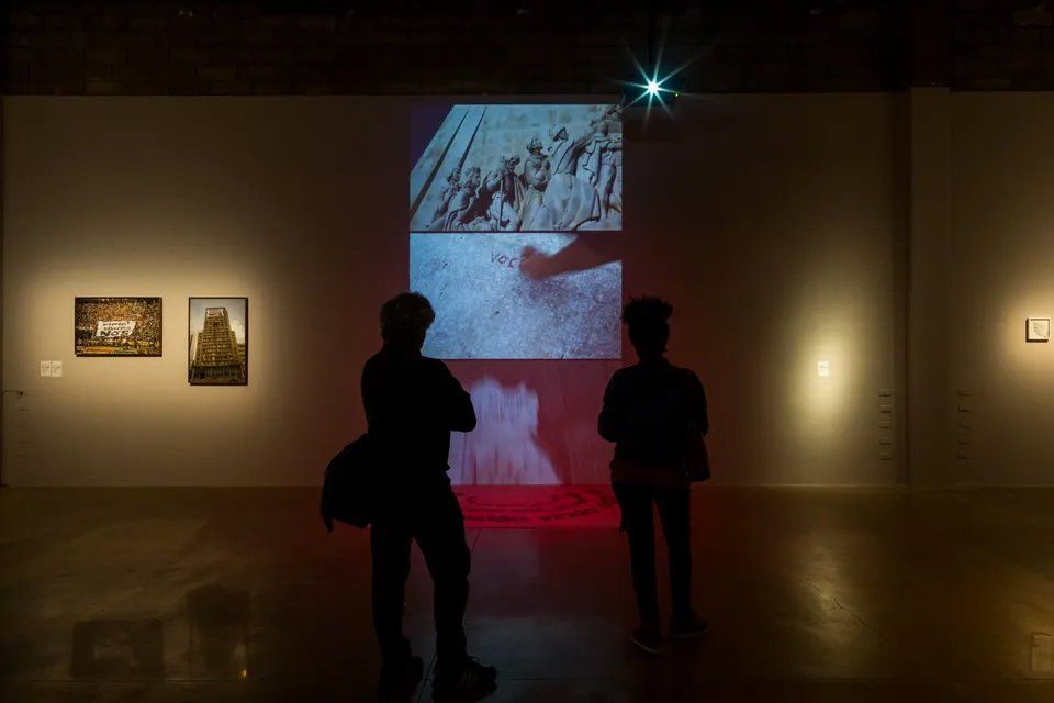 Duas pessoas estão diante de uma grande tela em uma sala escura.
