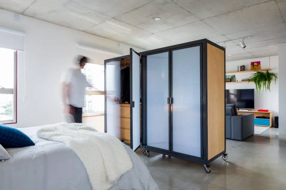 Um homem está parado em um quarto ao lado de uma cama e de um guarda-roupa.