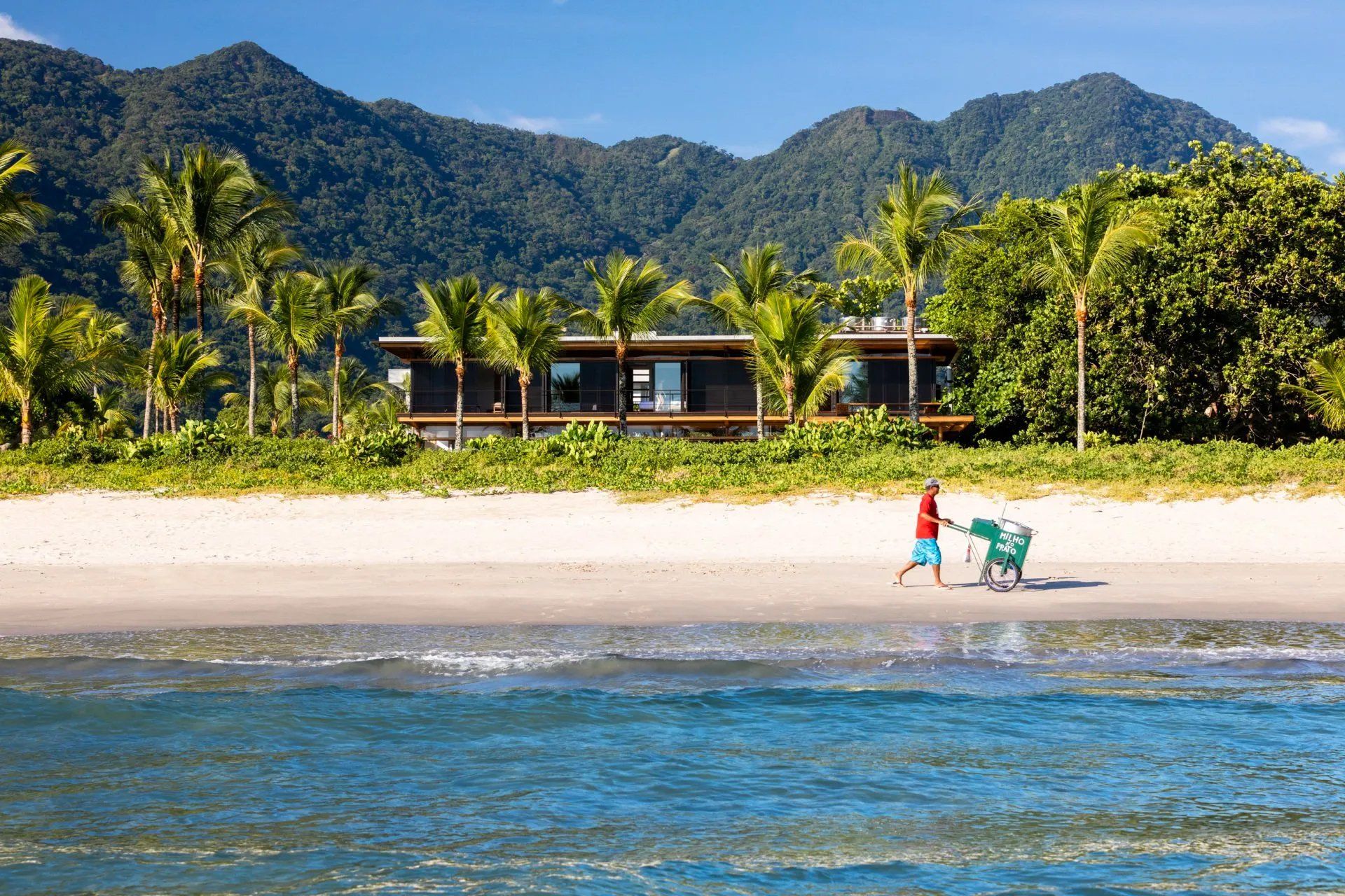 Um homem está empurrando um carrinho em uma praia com uma casa ao fundo.