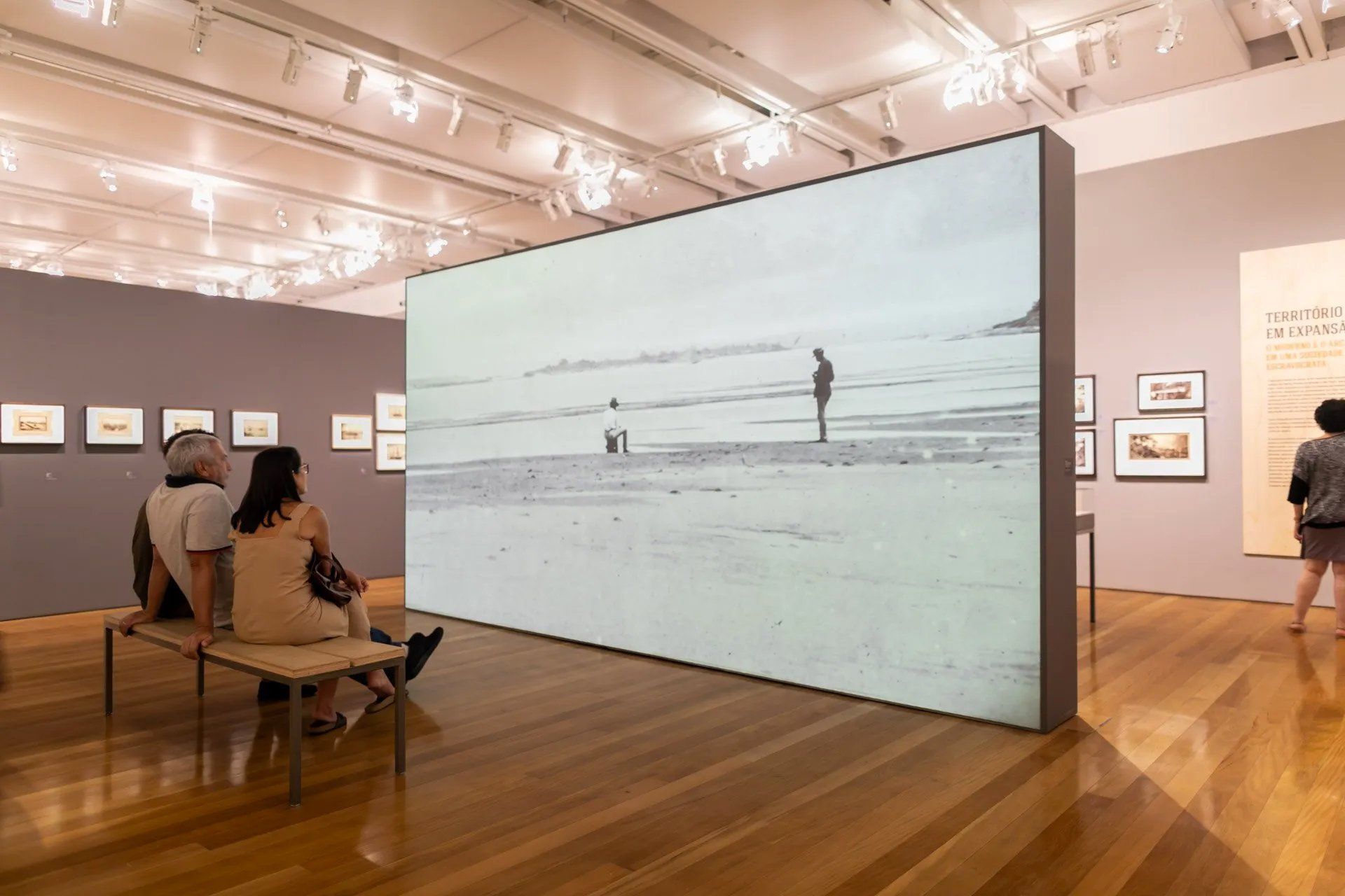 Algumas pessoas estão sentadas em um banco em frente a uma grande tela em um museu.
