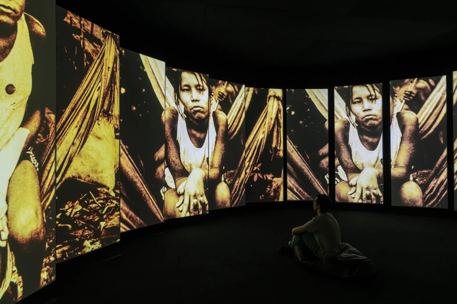 Uma pessoa está sentada em frente a uma parede de telas com fotos de pessoas nelas.