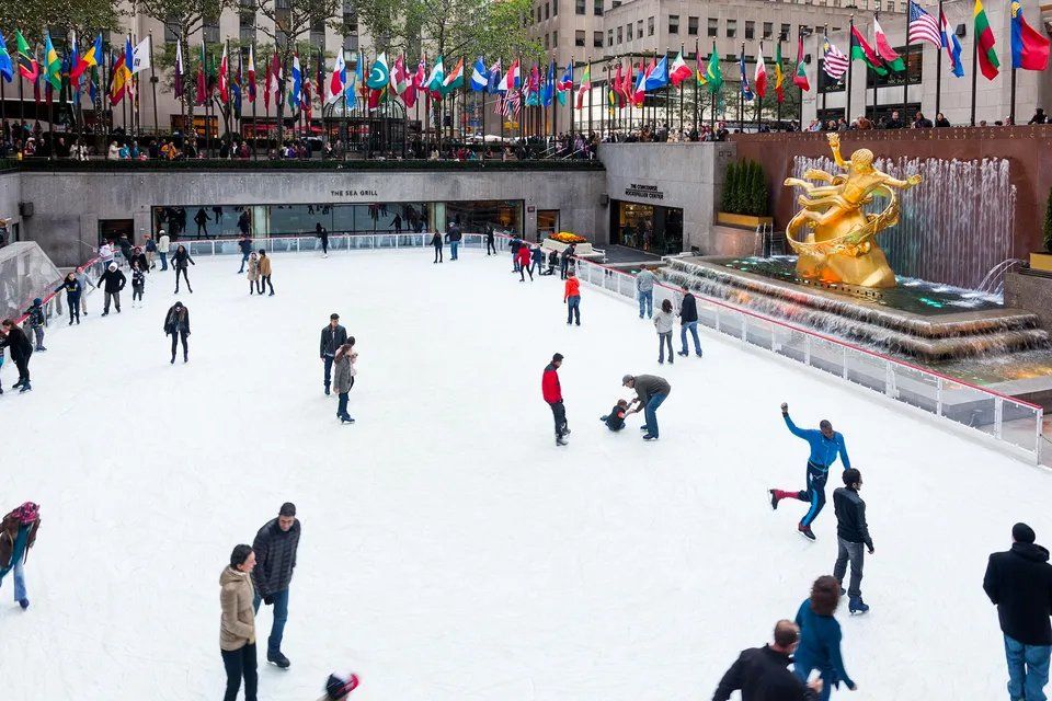 Um grupo de pessoas está patinando no gelo em uma pista em frente a uma fonte.