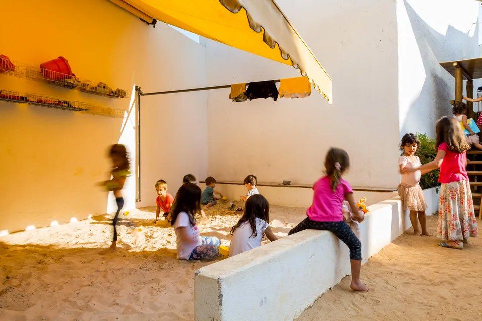 Um grupo de crianças brinca em uma caixa de areia sob um toldo.