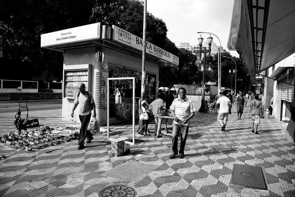Uma foto em preto e branco de pessoas andando na calçada em frente a um quiosque.
