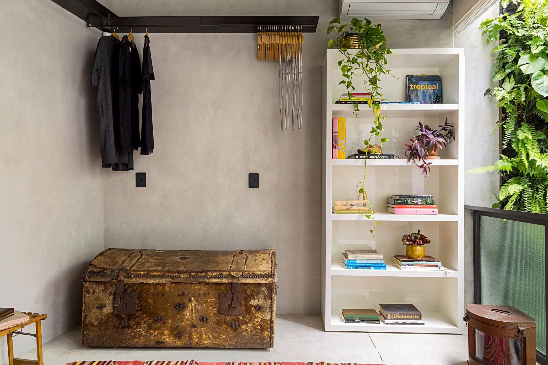 Um quarto com baú, prateleiras e roupas penduradas na parede.