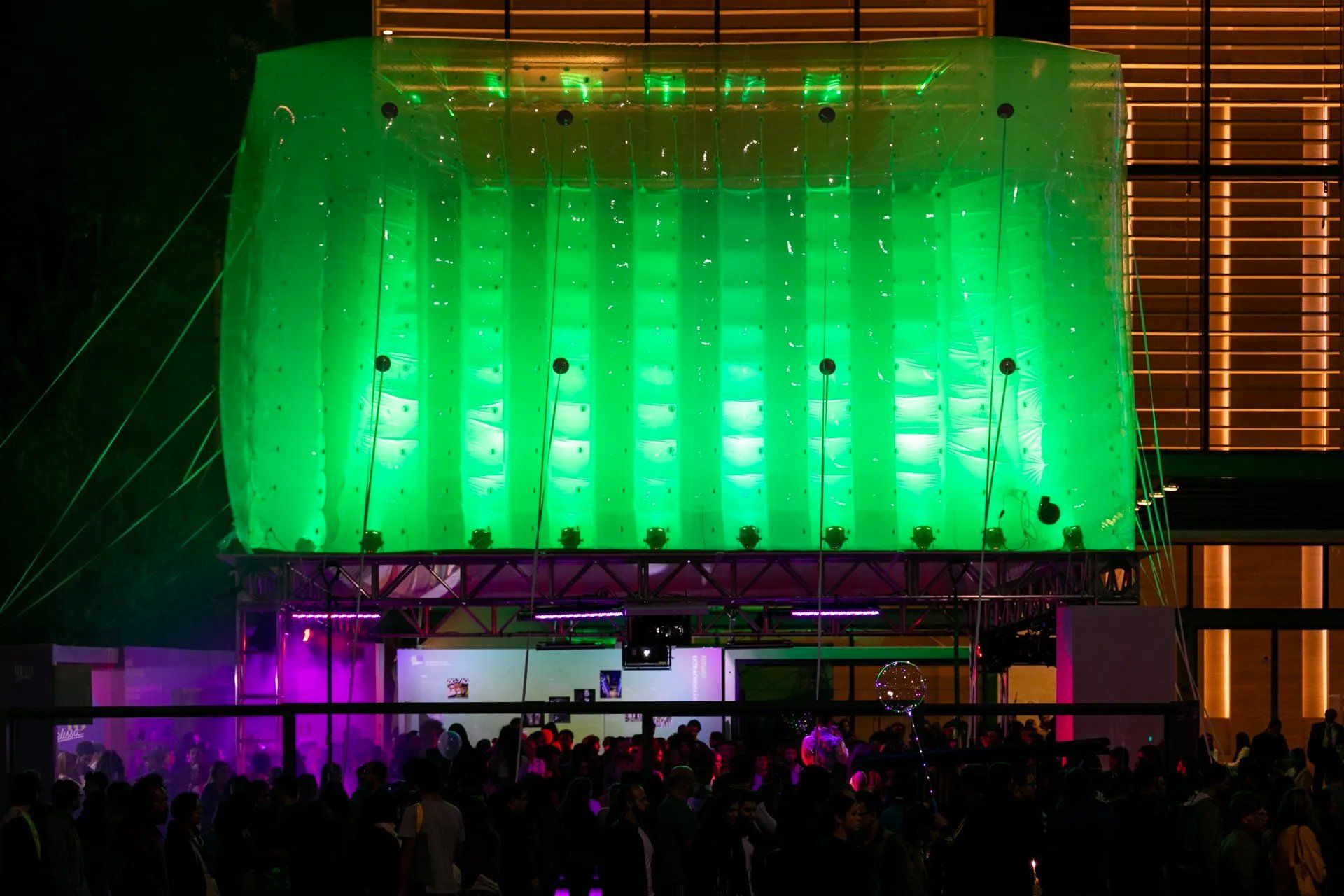 Uma multidão de pessoas está reunida em frente a um prédio com luzes verdes acesas.