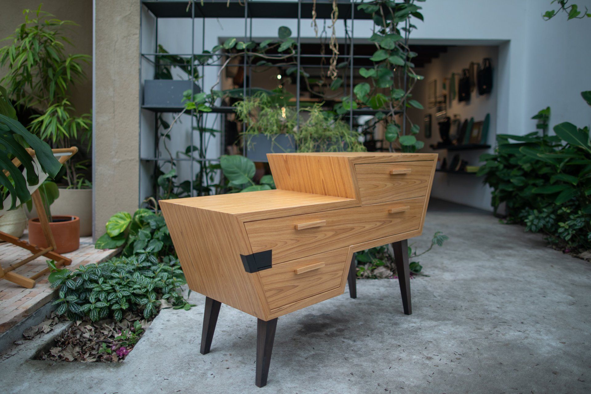 Uma cômoda de madeira está sobre um piso de concreto em um pátio cercado por plantas.