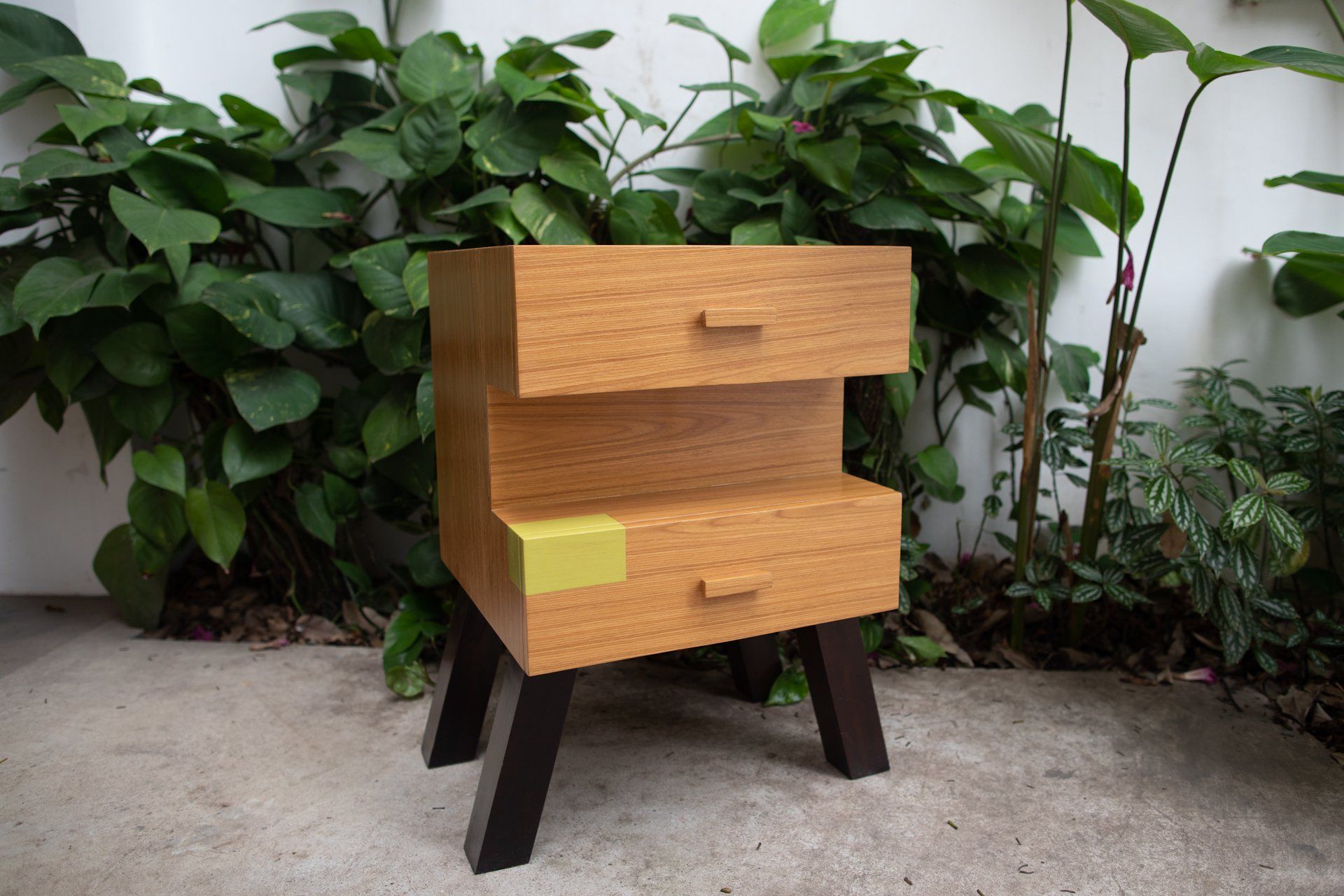 Uma pequena mesa de madeira com duas gavetas está em frente a um arbusto.