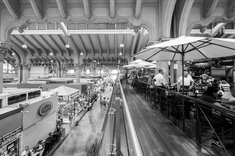 Uma foto em preto e branco de um mercado com pessoas sentadas em mesas sob guarda-chuvas.