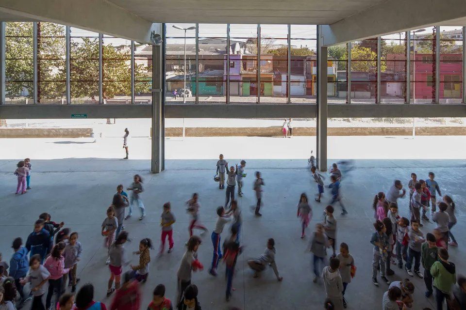 Um grupo de crianças brinca em uma sala grande com muitas janelas.