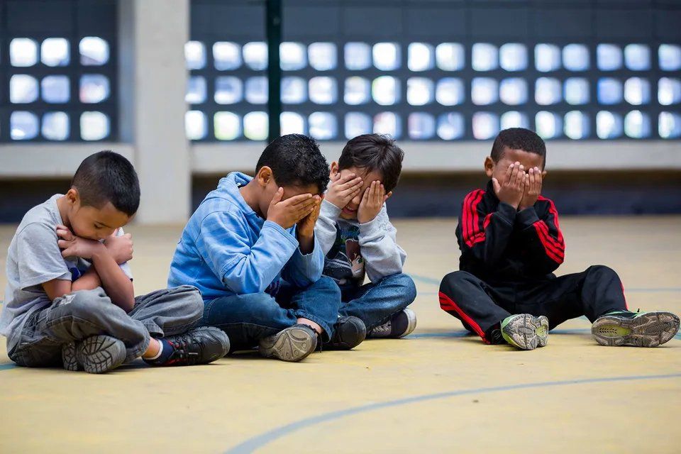 Um grupo de crianças está sentado no chão cobrindo o rosto.