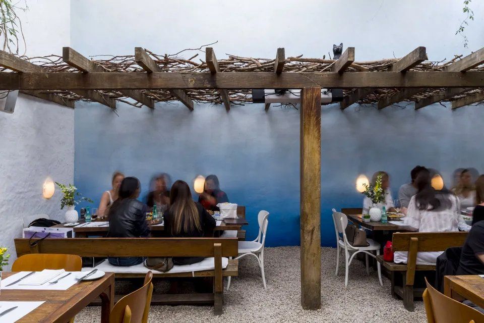 Um grupo de pessoas está sentado às mesas de um restaurante sob uma pérgula.