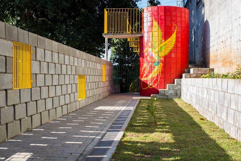 Uma parede de tijolos com janelas amarelas e um cilindro vermelho ao fundo.