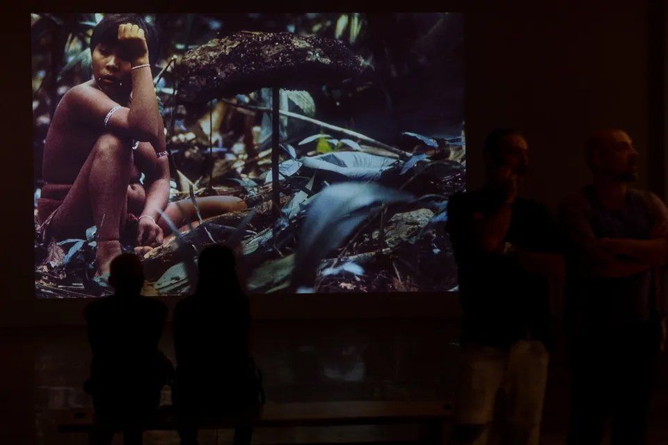 Um grupo de pessoas está diante de uma grande tela com a foto de um homem.