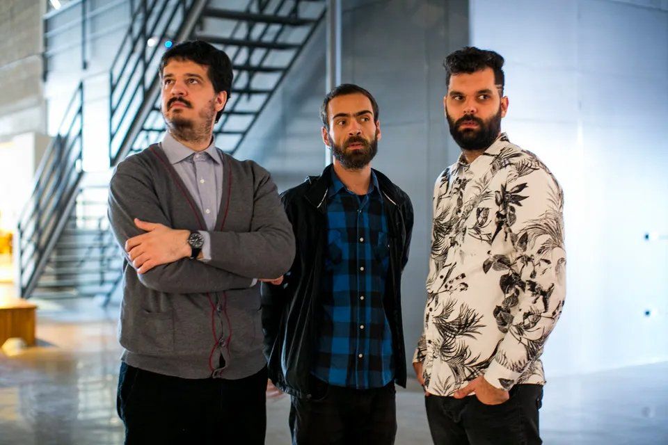 Três homens barbudos estão parados um ao lado do outro em frente a uma escada.