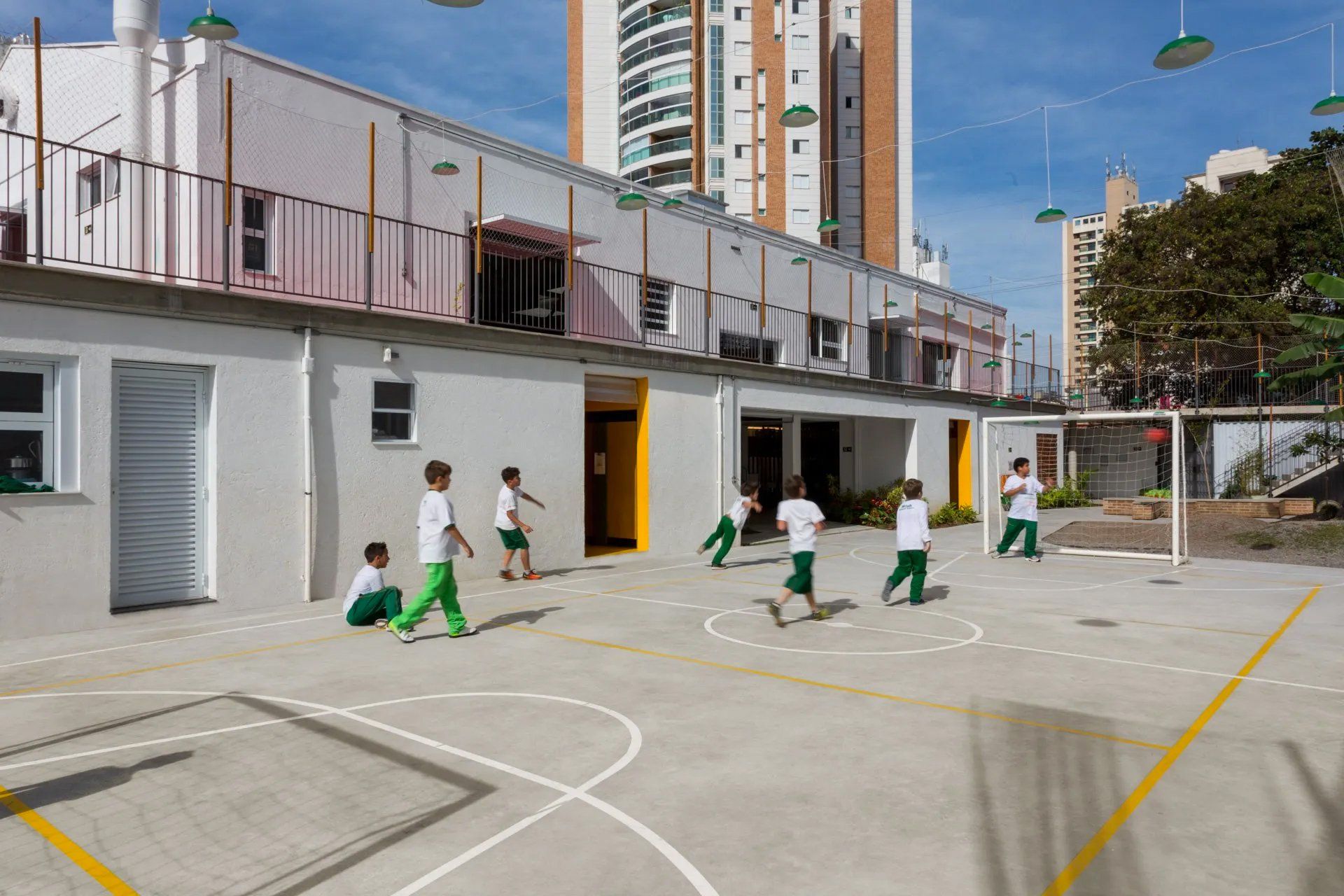 Um grupo de crianças joga futebol em uma quadra em frente a um prédio.