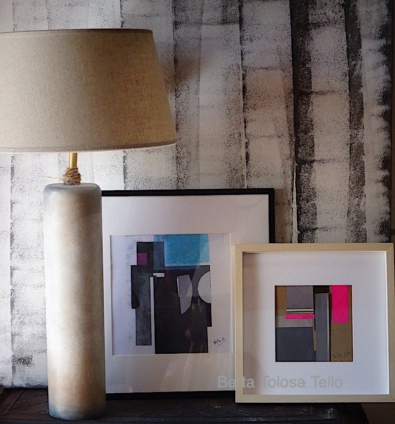 Una lámpara está sobre una mesa junto a dos cuadros enmarcados de Bianca Coloba Tello.