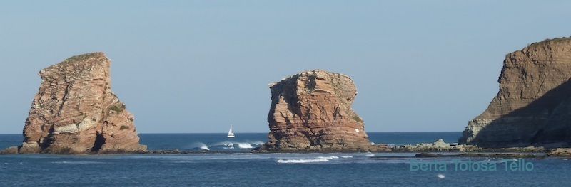 Tres rocas en el océano con un velero en la distancia