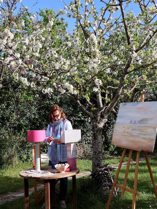 Una mujer está sentada en una mesa con una lámpara y un cuadro en un caballete.