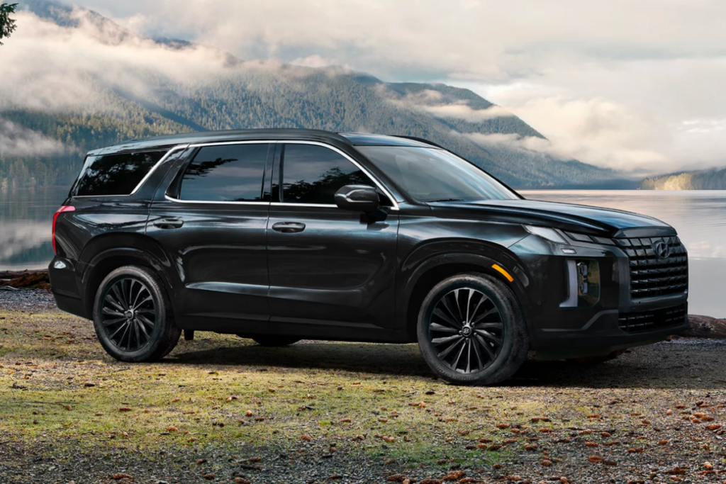 A black hyundai palisade is parked next to a lake with mountains in the background.