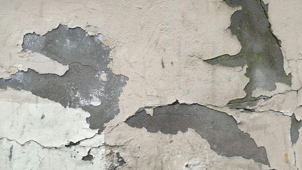 Cracked, peeling beige paint on a gray concrete wall, revealing the underlying surface.