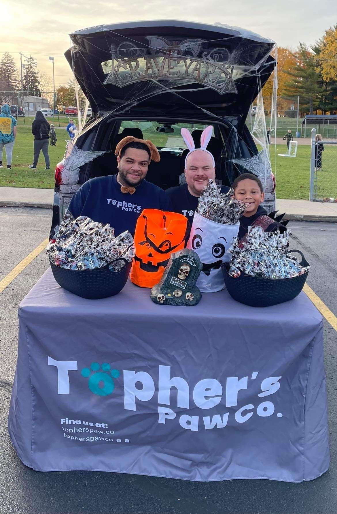 A group of people are sitting at a table in front of a car trunk.