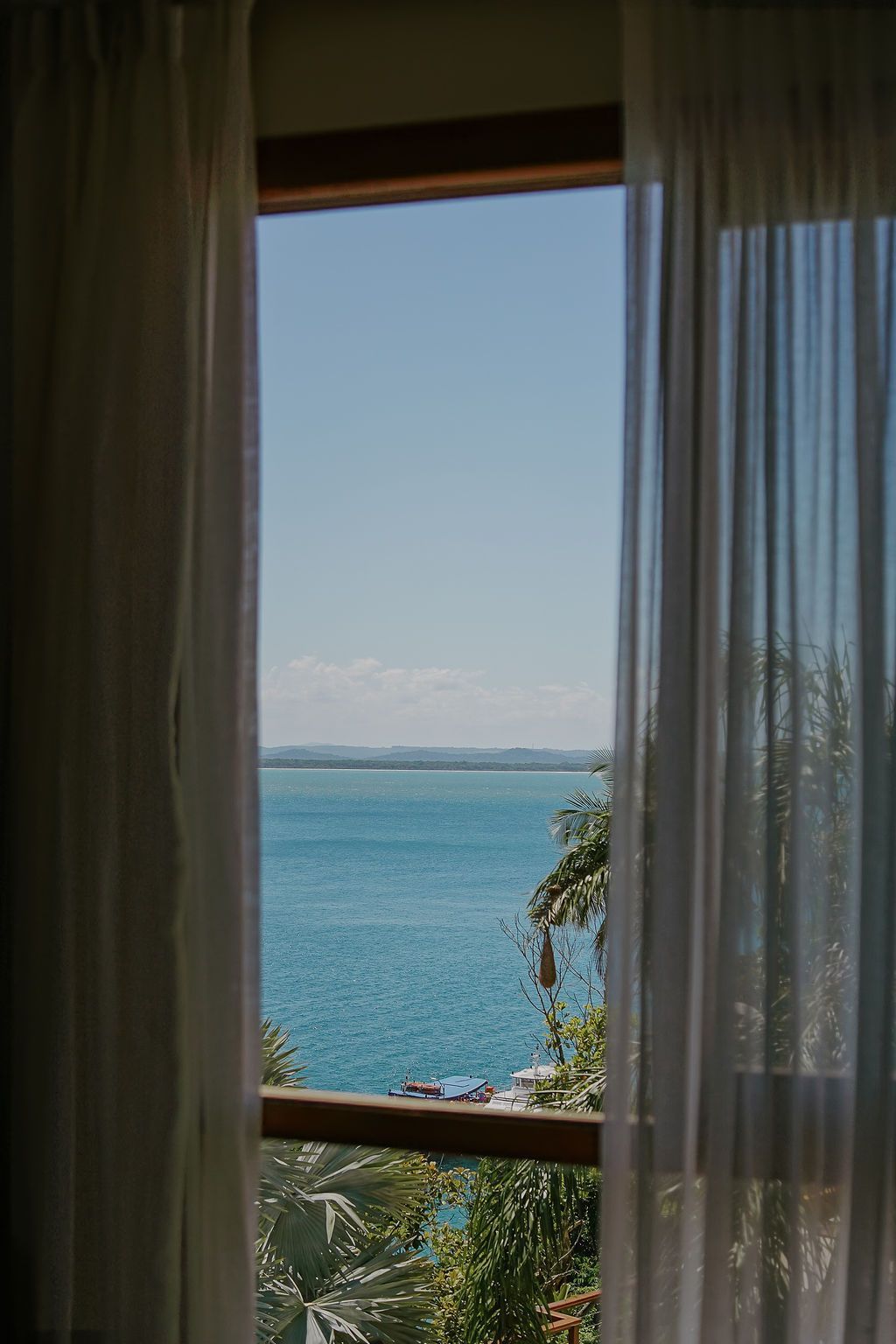 Vista da janela para o oceano e o céu, emoldurada por cortinas brancas, com vegetação abaixo.