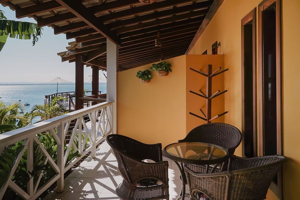 Varanda com vista para o mar, móveis de vime, paredes amarelas, detalhes em madeira e plantas.