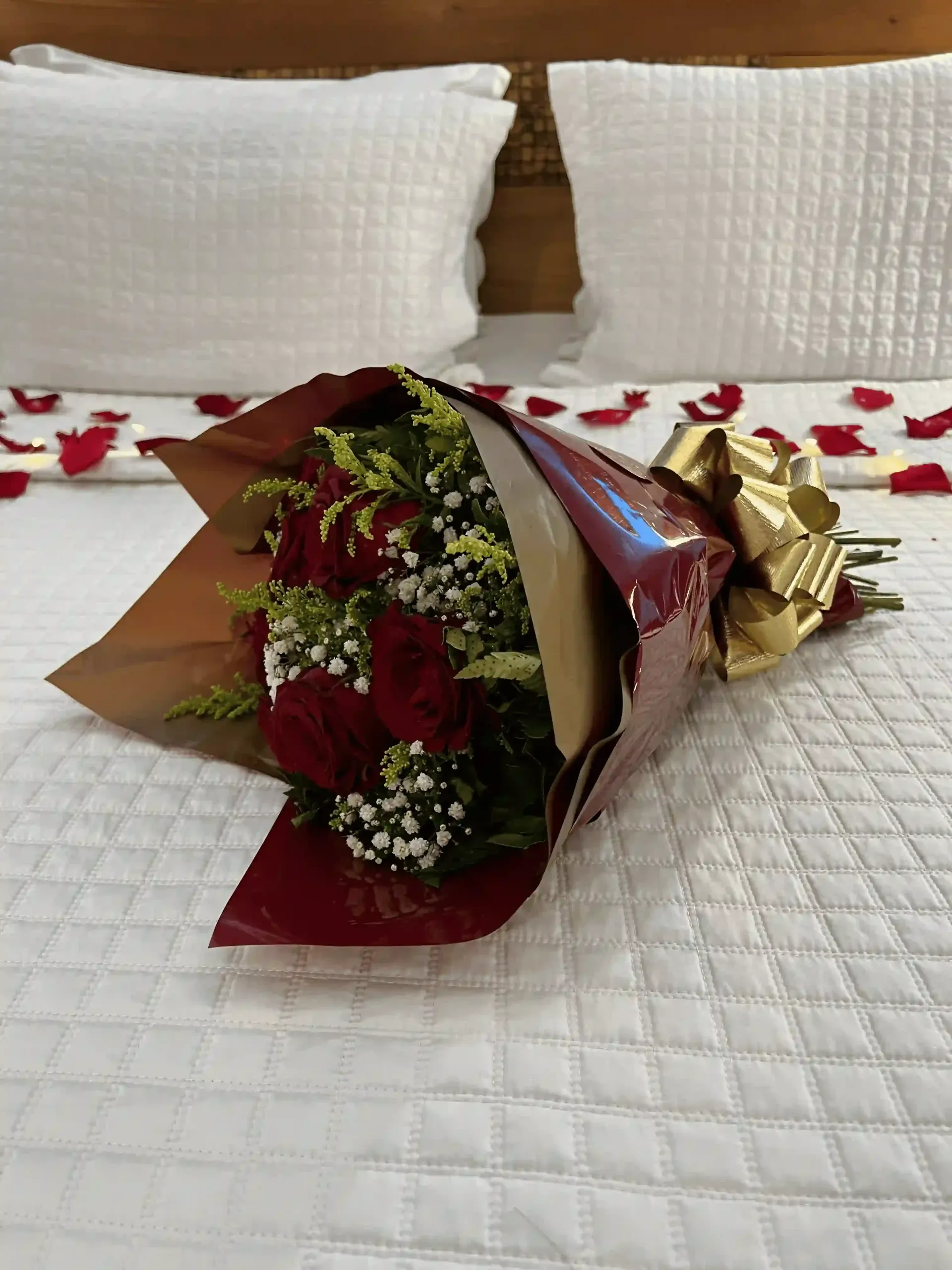 Buquê de rosas vermelhas embrulhadas em papel bordô e fita dourada sobre uma cama com pétalas de rosas espalhadas.