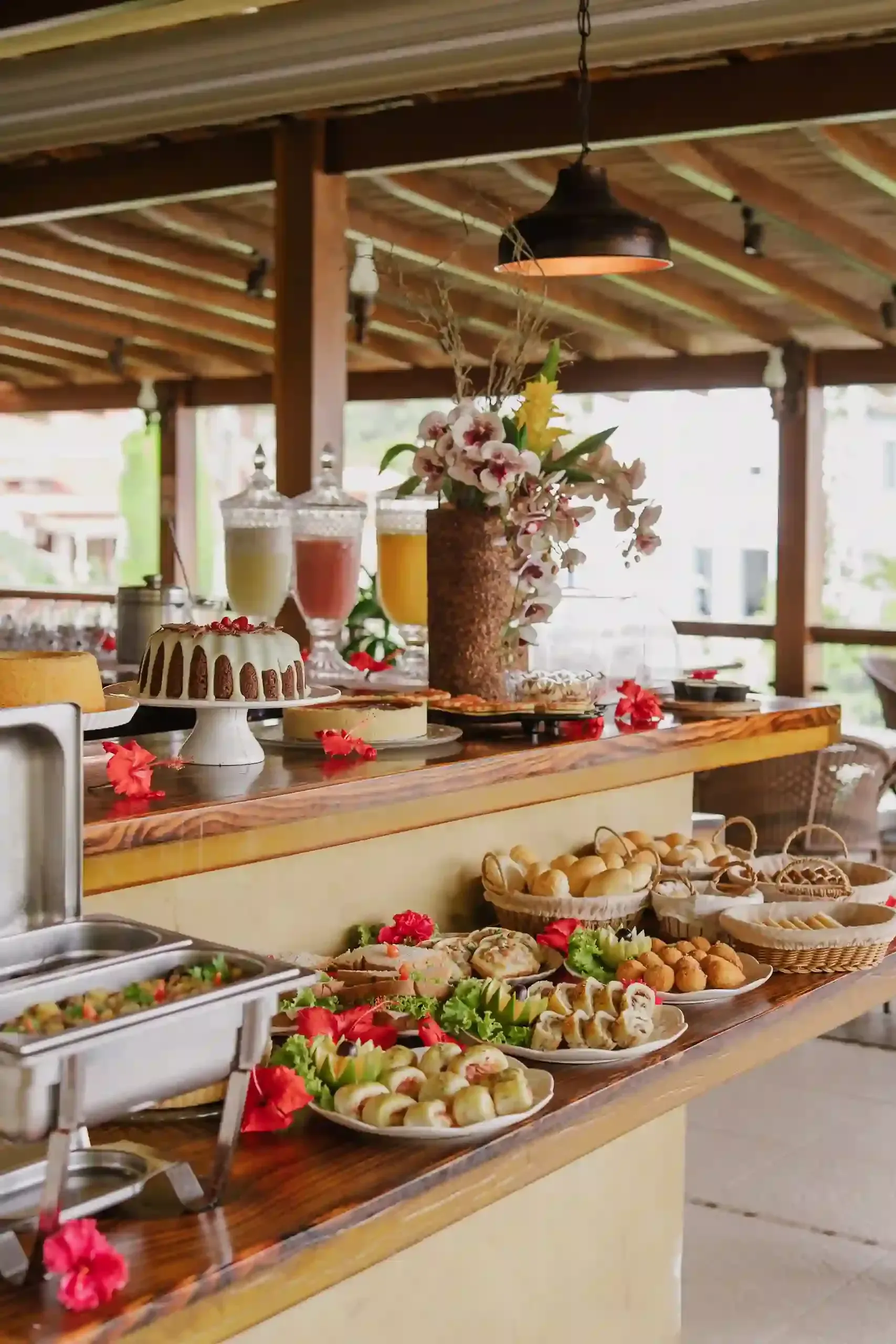 Mesa de bufê com comidas, bebidas e decoração floral. Ambiente externo com vigas de madeira.