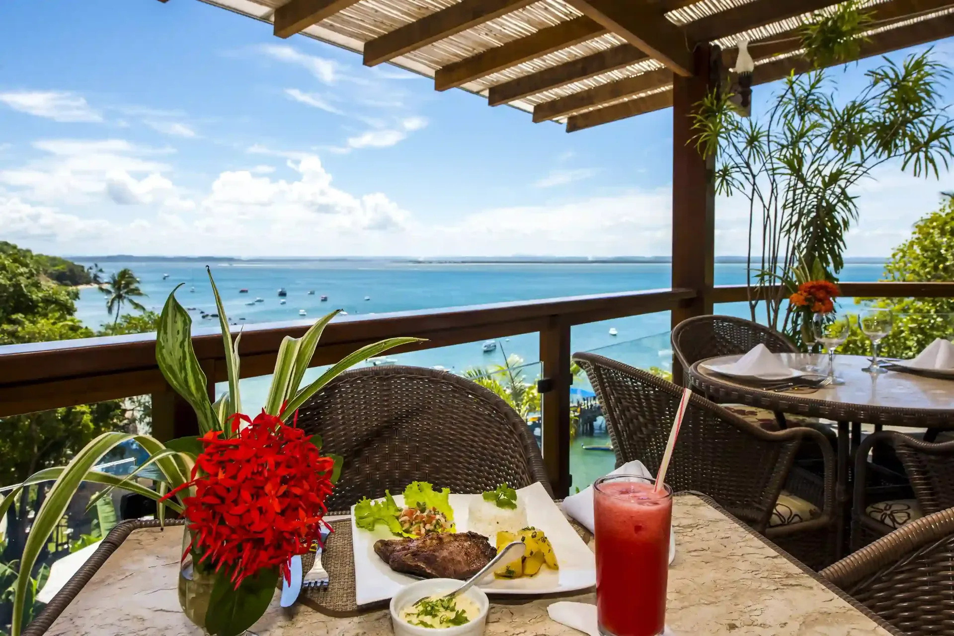 Mesa de restaurante com comida e bebida, com vista para o mar em um dia ensolarado.