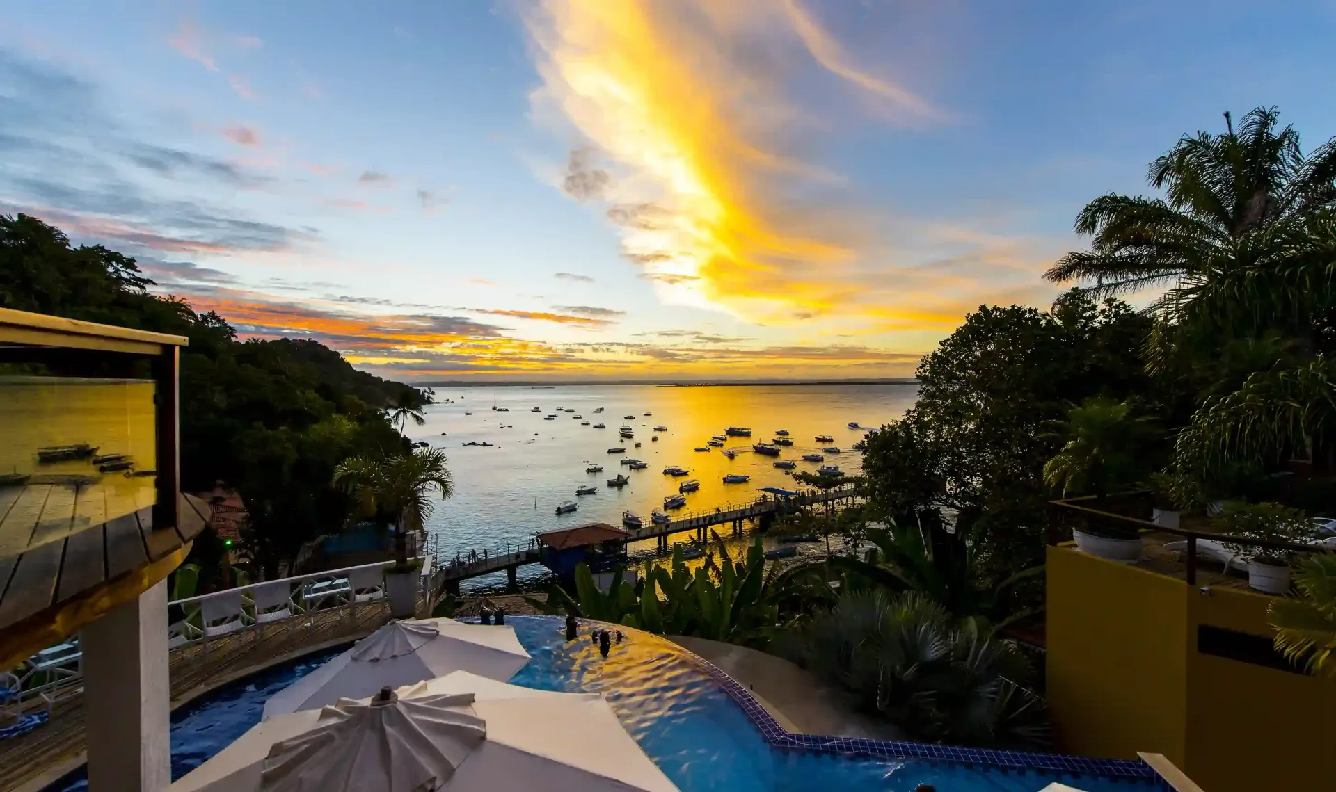 Pôr do sol sobre a baía com barcos, visto de uma piscina. Céu dourado, água azul, árvores exuberantes.