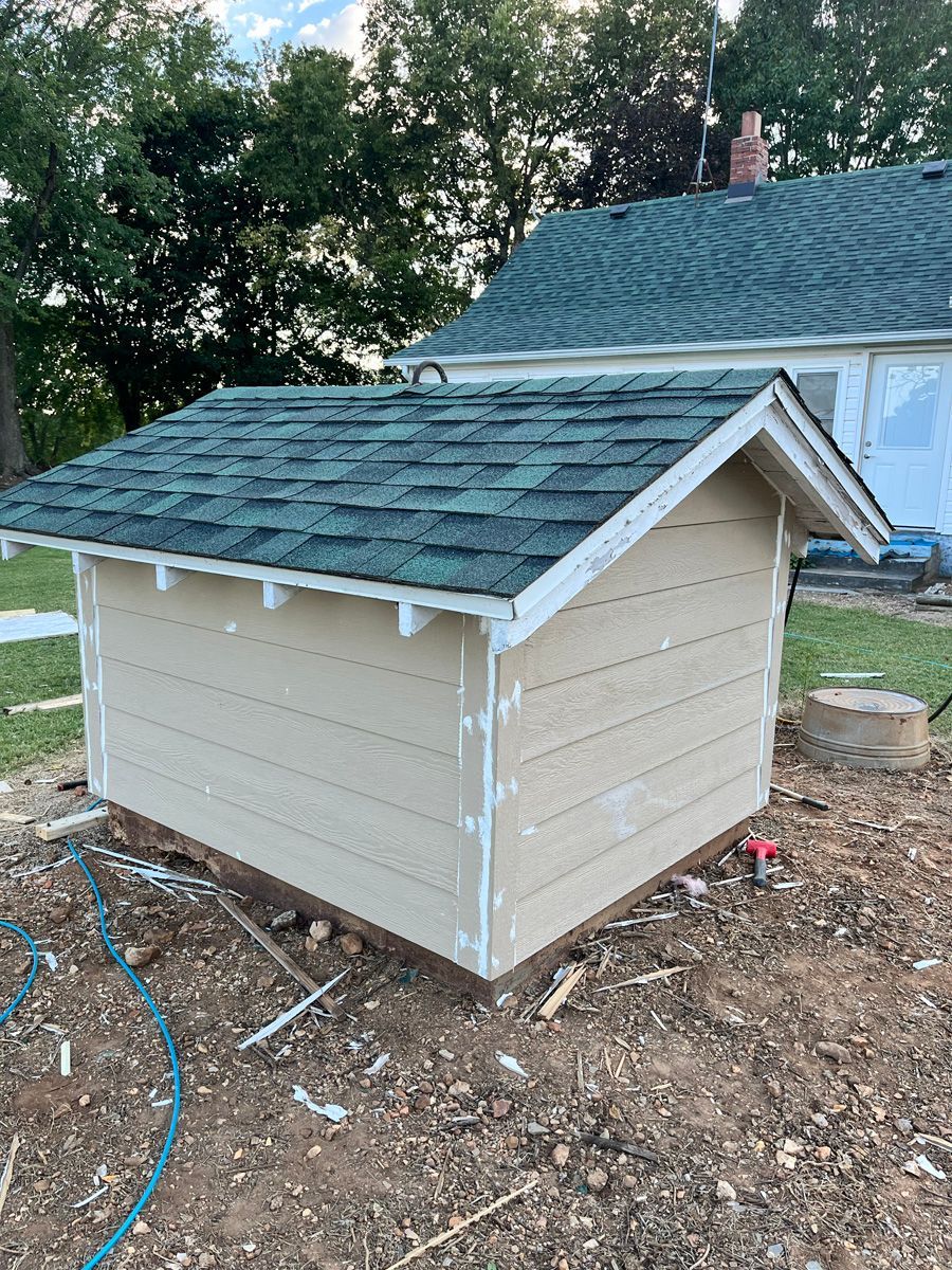 A small dog house with a blue roof is sitting in the dirt in front of a house.