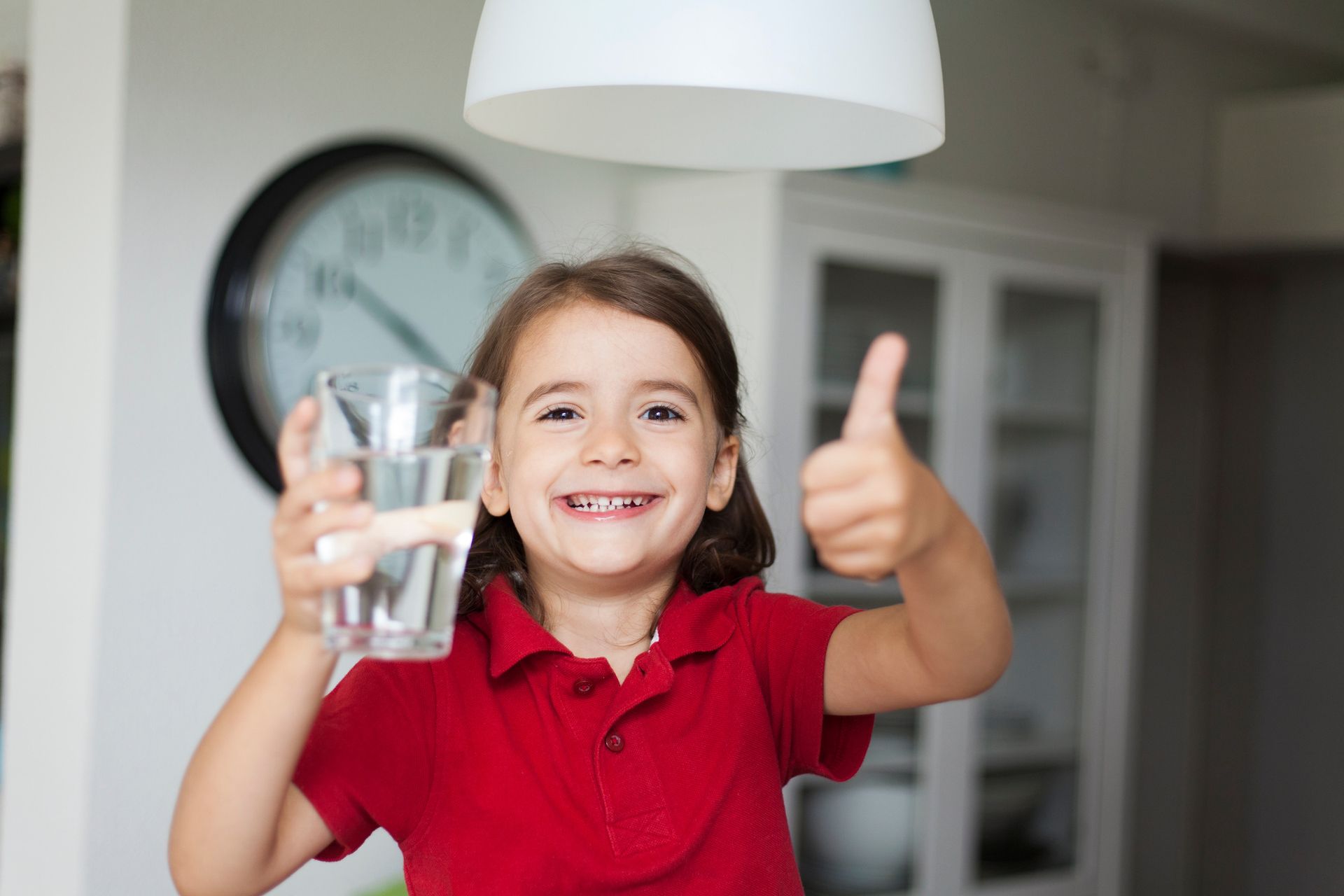 A little girl is holding a glass of water and giving a thumbs up.