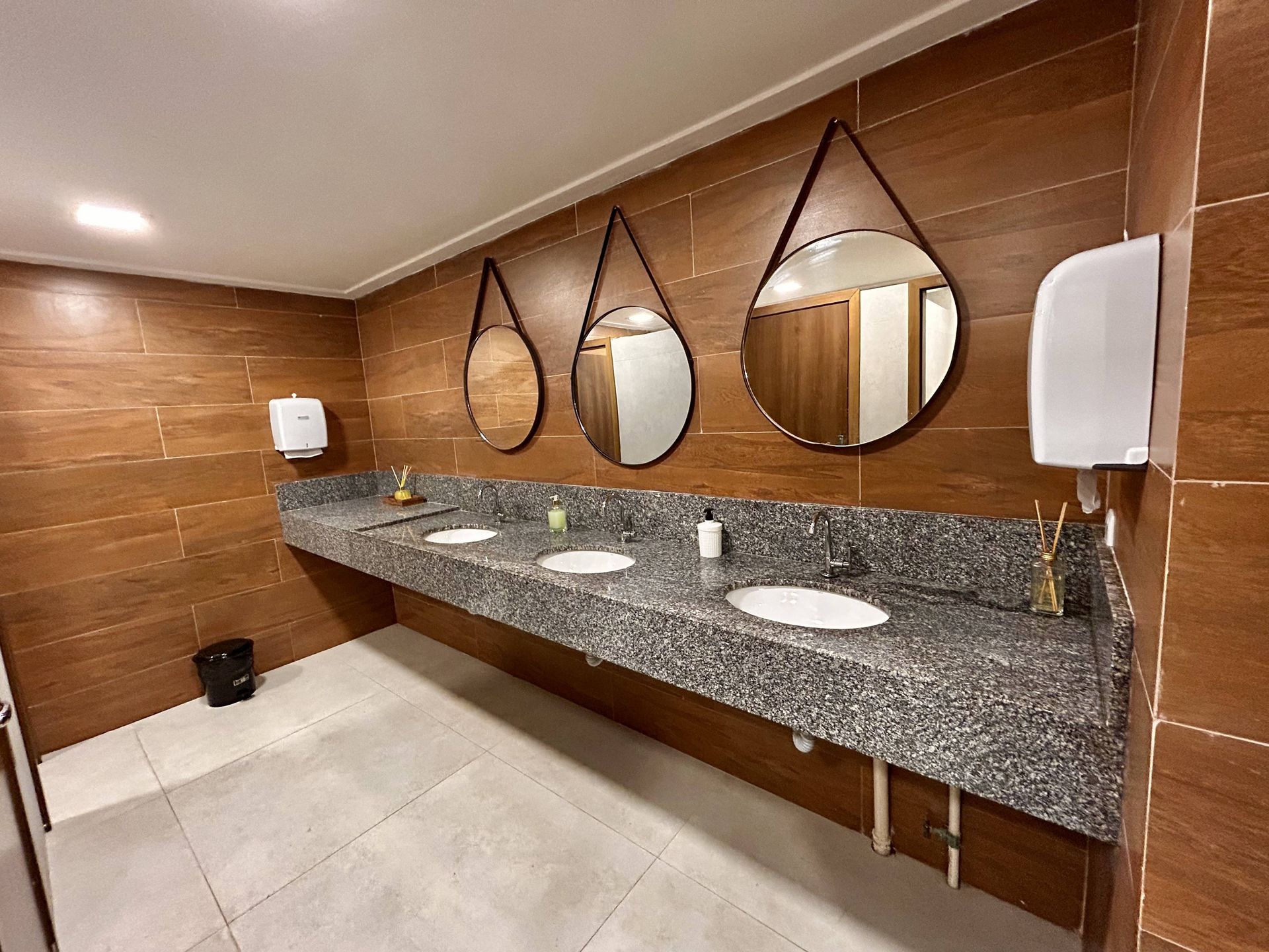 Banheiro público com três pias, espelhos em formato de gota, paredes revestidas com azulejos marrons e bancada de granito.